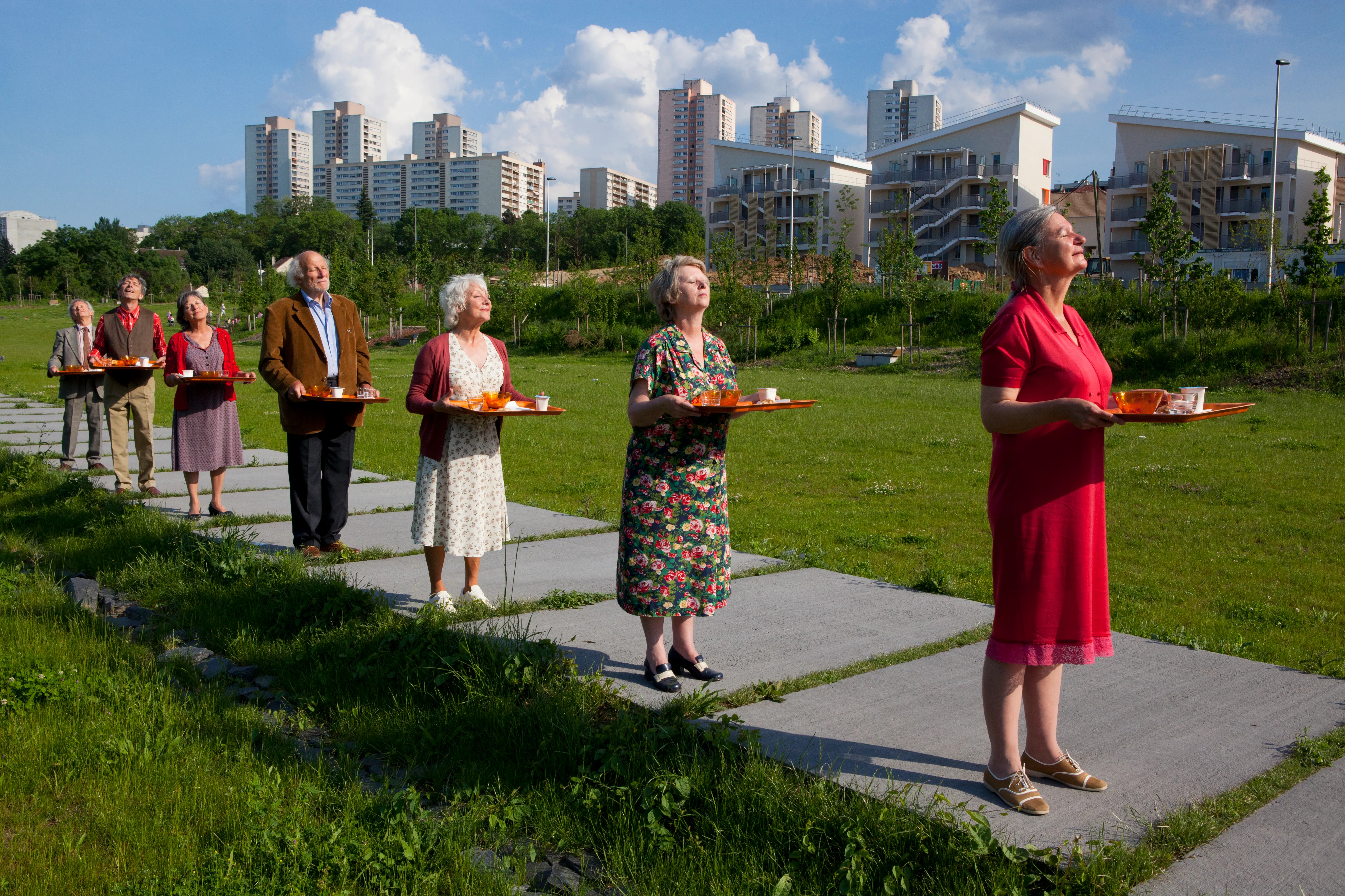 Line of elderly people on a conctrete path surrounded by grass, part of a performance at the Bradford Festival in July.