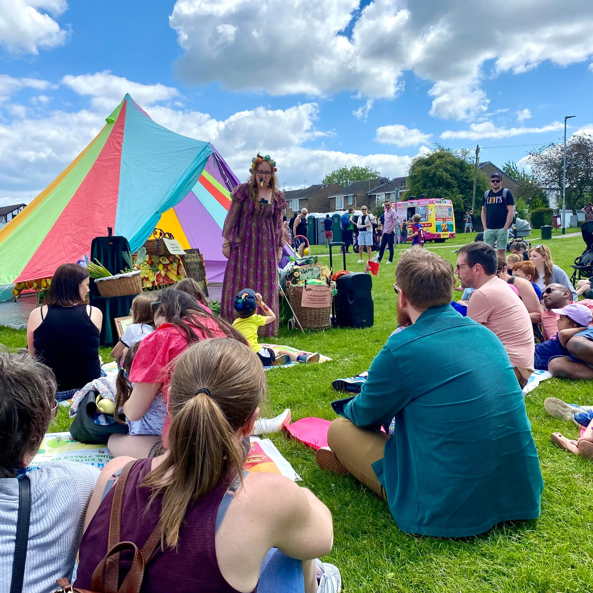 People sitting on the grass on a sunny day at Farsley Festival listening to an entertainer.