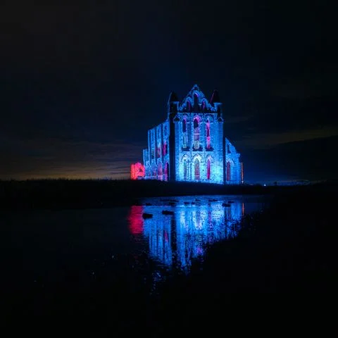 Illuminated Gothic ruins of Whitby Abbey lit up in vibrant colors after dark during October Halloween season