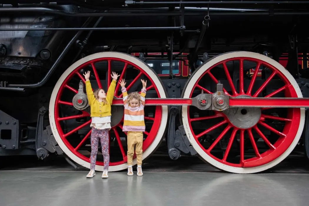 two children with arms stretched up trying to make themselves as tall as one of the train wheels in the NAtional Railway Museum in York, part of the Summer Holiday in a Day event. 