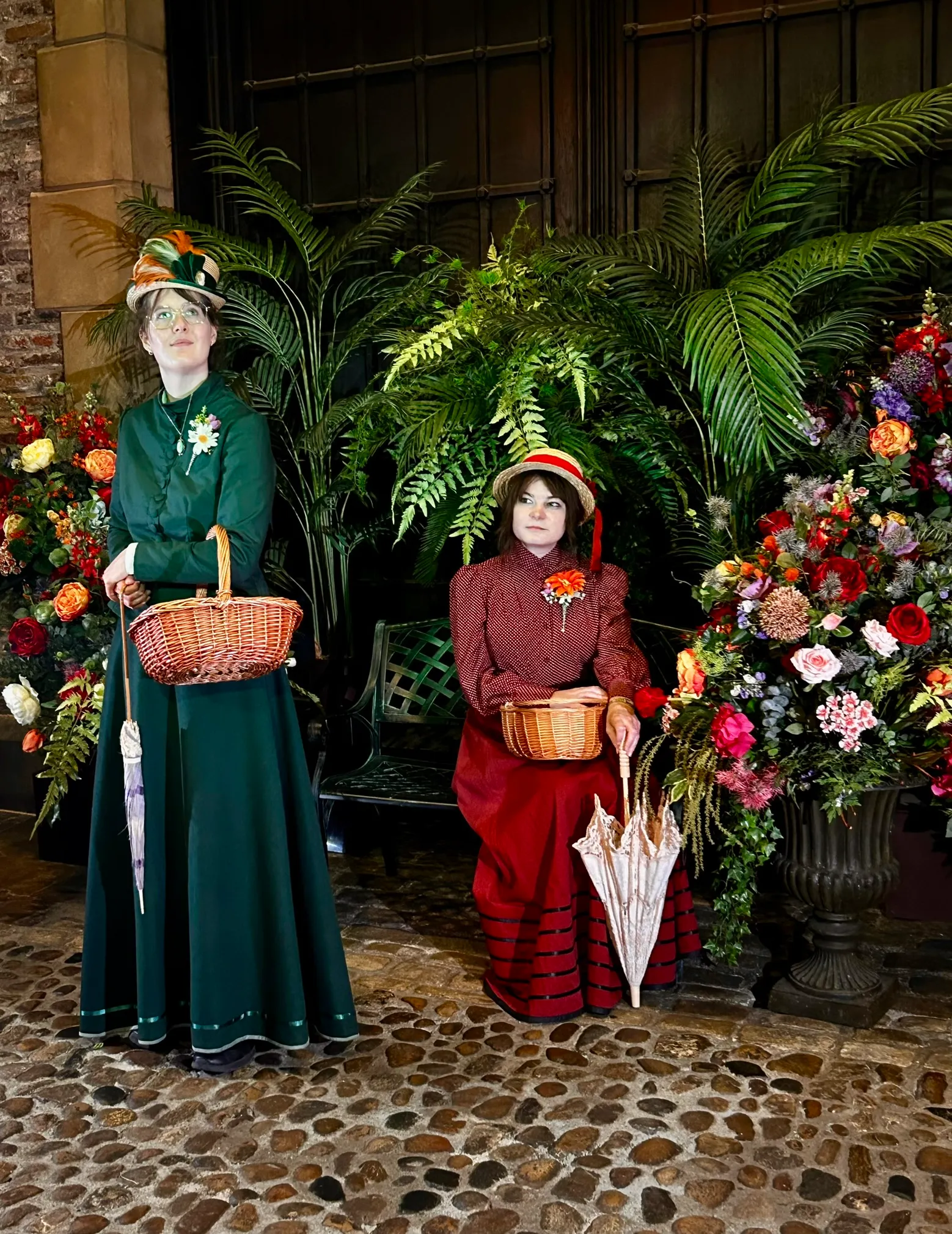 Two ladies in Victorian attire in front of a beautiful flower display