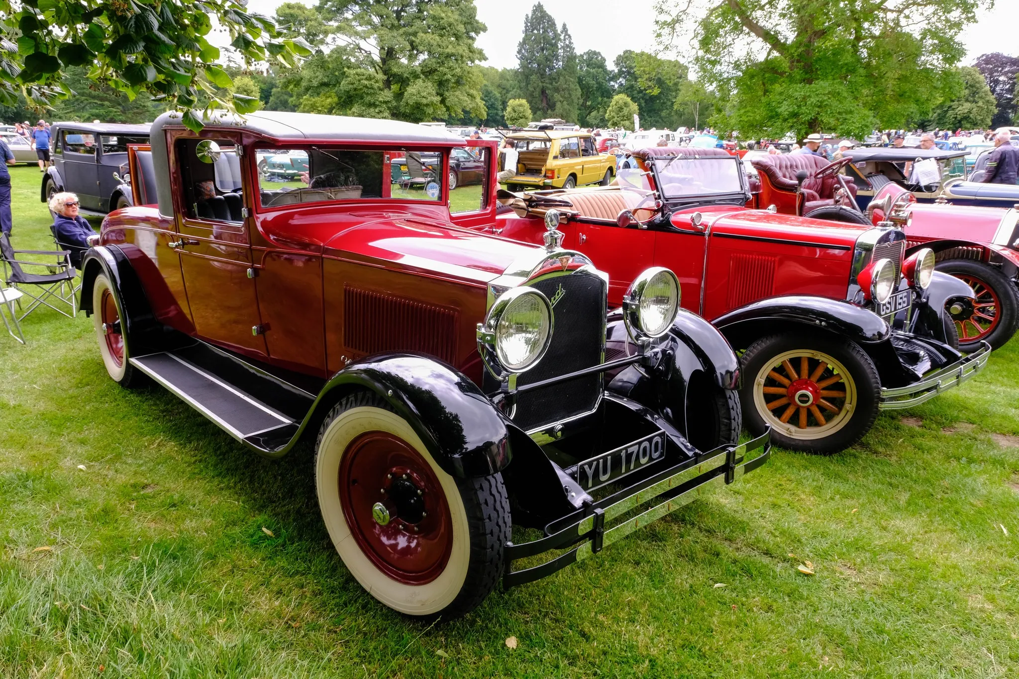 Red historic car in parkland with other cars in the background